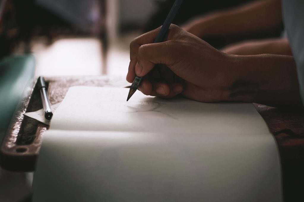 pexels-photo-5299834-5299834 A close-up shot of a hand sketching art on paper using a pencil, in a dimly lit space.